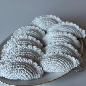 May include: A collection of white, crocheted, half-circle shaped items with a scalloped edge, arranged on a white and gold-rimmed plate. The handmade items appear to be coasters or decorative pieces, showcasing a textured, detailed stitch.