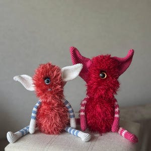 May include: Two handmade crochet monster dolls. One is red with blue and white striped arms and legs, white ears, and a blue eye. The other is a darker red with pink ears and a yellow eye. Both have fluffy bodies and are sitting.