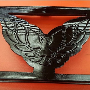May include: Black metal emblem with a stylized bird design. The bird has a wingspan and a raised head. The emblem is rectangular with a smooth, glossy finish.