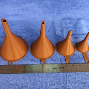 May include: Four orange plastic funnels of different sizes lined up on a blue surface with a ruler underneath. The ruler is marked in inches and has the words "Stainless Steel" and "Westcott" printed on it.