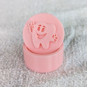 May include: A pink plastic tooth-shaped box with a smiling face and a toothbrush in its hand. The box is closed and has a lid.