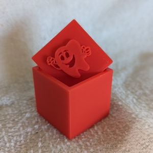 以下が含まれることがあります： 赤い立方体の箱で、取り外し可能な蓋が付いています。蓋には、腕と脚のある笑顔の漫画の歯の切り抜きがあります。