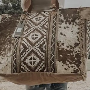 May include: Large brown and white cowhide tote bag with a geometric patterned center panel. The bag has two handles and a tag attached. The bag is made of a combination of materials, including cowhide and woven fabric.