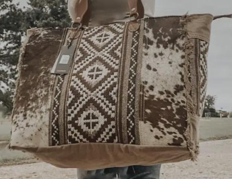 May include: Large brown and white cowhide tote bag with a geometric patterned center panel. The bag has two handles and a tag attached. The bag is made of a combination of materials, including cowhide and woven fabric.
