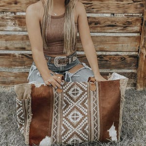 May include: A large brown and white tote bag with a geometric pattern and leather handles. The bag has a cowhide texture and a fringed edge. The bag is sitting on the grass.