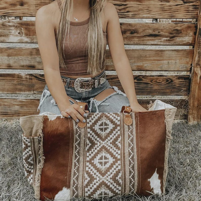May include: A large brown and white tote bag with a geometric pattern and leather handles. The bag has a cowhide texture and a fringed edge. The bag is sitting on the grass.