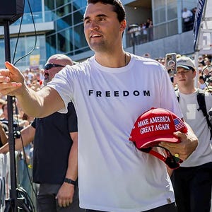 May include: A white t-shirt with the word "FREEDOM" printed in black. A red baseball cap with the text "MAKE AMERICA GREAT AGAIN" is held in the hand. The cap has an American flag design.