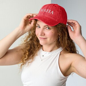 May include: A woman with curly red hair is wearing a red baseball cap with the word "MAHA" embroidered on the front in white. She is wearing a white tank top and has a silver necklace on.
