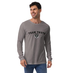 Può includere: Una maglietta grigia a maniche lunghe con la scritta "TEAM TRUTH" in lettere nere sopra un emblema circolare. L'emblema presenta un disegno a scudo con le lettere "HB" e uno stendardo.
