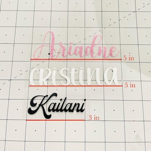 May include: Three names, "Ariadne", "Cristina", and "Kailani", are written in different styles and colors on a white background with a grid pattern. The names are 5 inches wide and the bottom name is 3 inches wide.