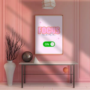 May include: A framed print with the words "FOCUS mode ON" in pink and green. The print is displayed on a white table with a vase, a decorative sphere, and a geometric shape. The background is a pink wall.