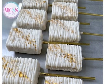 May include: A row of white chocolate covered treats with gold sprinkles on top, arranged on a gray surface. Each treat is on a gold stick.