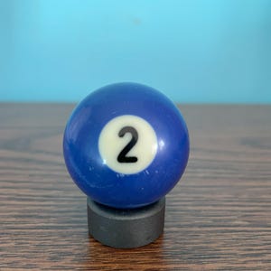 Peut inclure: Une boule de billard bleue avec le numéro 2 blanc dessus. La boule est posée sur un support noir.