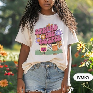 以下が含まれることがあります： 福音を伝える2つの瓶のレトロなグラフィックが描かれた白いTシャツ。瓶は脚と腕と顔を持っています。シャツのテキストは「Spread the Gospel」です。