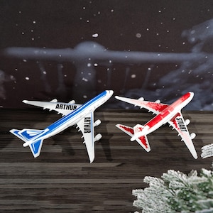 May include: Two toy airplanes, one blue and white with "ARTHUR" printed on the side, and another red and white with "STUART" printed on the side. The planes are on a dark wood surface with a snowy background.