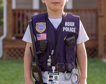 May include: A dark blue police vest featuring the text NOAH POLICE, a gold badge, an American flag patch, a walkie-talkie, a whistle, a black holster, and silver handcuffs. A white t-shirt is worn underneath, and a police officer ID card is attached.
