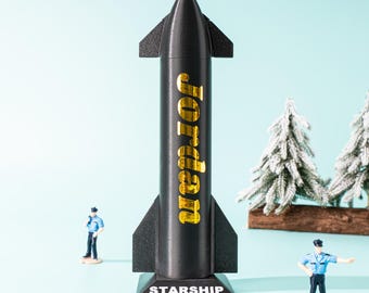 May include: A black Starship model with gold lettering reading "Jordan". The model has fins and sits on a base labelled "STARSHIP". Two miniature police officer figures and two miniature Christmas trees are also present in the image.