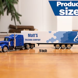 May include: Blue toy semi-truck with a white trailer. The trailer has a blue and white geometric pattern and the text "Matt's Trucking Company". The truck is 23 cm long and 3.8 cm tall.