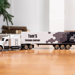 May include: White toy semi-truck with a trailer. The trailer has a black and white design with the text "Tom's Trucking Company".
