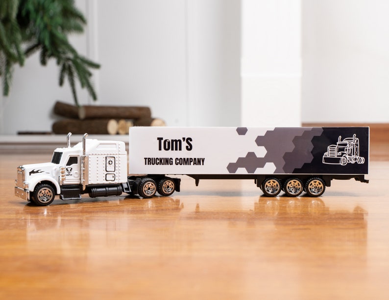May include: White toy semi-truck with a trailer. The trailer has a black and white design with the text "Tom's Trucking Company".