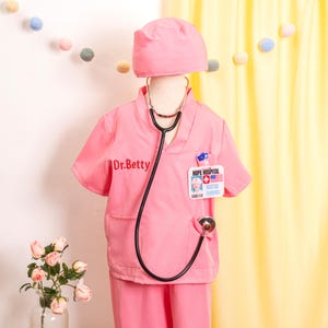 May include: A pink doctor's costume set, including a scrub top with "Dr. Betty" embroidered on the front, matching pants, a cap, and a stethoscope. The scrub top has a "Hope Hospital" name tag. A fun outfit for dress-up play.