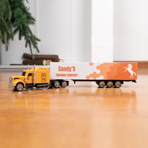 May include: A yellow toy semi-truck with a white trailer. The trailer has an orange and white design with the text "Sandy's Trucking Company" and a white horse silhouette.