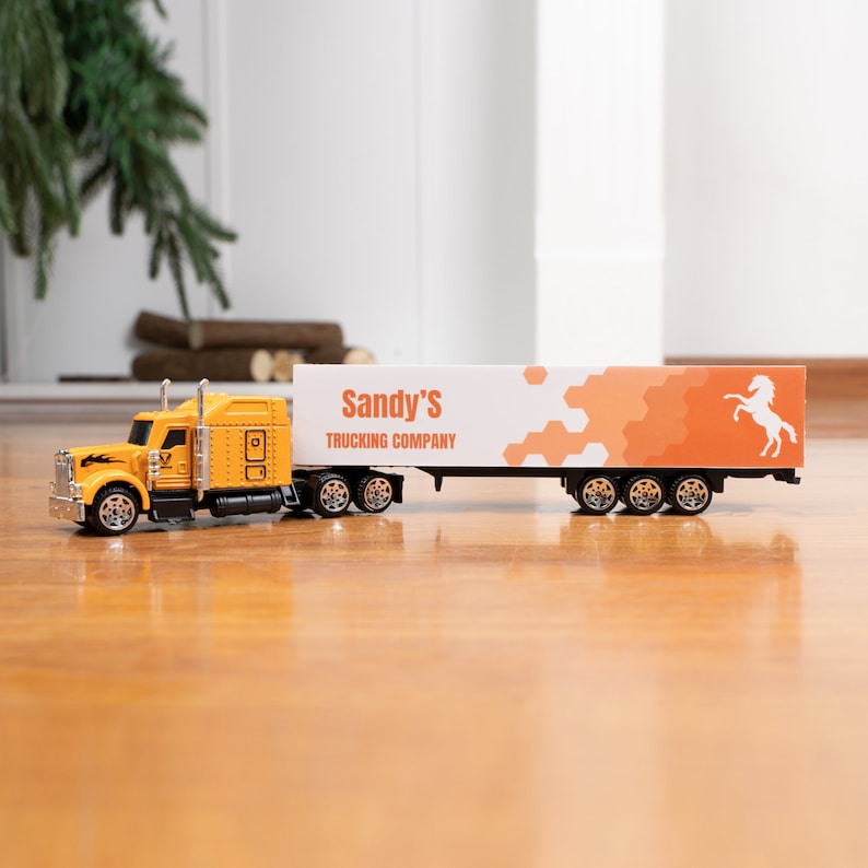 May include: A yellow toy semi-truck with a white trailer. The trailer has an orange and white design with the text "Sandy's Trucking Company" and a white horse silhouette.
