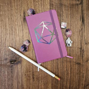 May include: A purple notebook with a geometric design, a white pencil, dice, and a miniature figurine are arranged on a wooden surface. The notebook has a matching ribbon bookmark. The dice are translucent with black markings.
