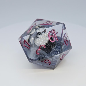 May include: A clear, multi-sided die with pink numbers and a black and white interior design. The die contains a cluster of white, pearl-like beads. The numbers are in a pink font.