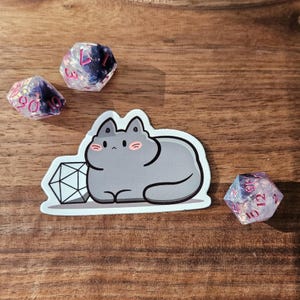 May include: A gray cat sticker with a D20 die and three other multi-sided dice. The dice are clear with pink numbers and glitter. The sticker and dice are on a wooden surface. The cat sticker has pink cheeks.