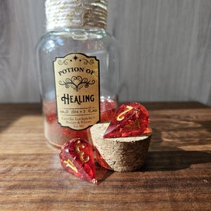 Puede incluir: Una botella de vidrio etiquetada como "Potion of Healing" con purpurina roja en su interior, junto a dos dados de resina roja con números dorados. La botella tiene un tapón de corcho y está decorada con hilo. Los dados tienen forma de lágrima.
