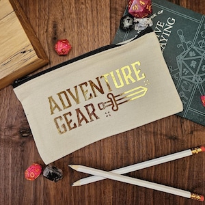May include: A beige canvas pencil case with the words "ADVENTURE GEAR" in gold lettering and a sword graphic. The case has a black zip and is on a wooden surface with pencils, dice, and a book.