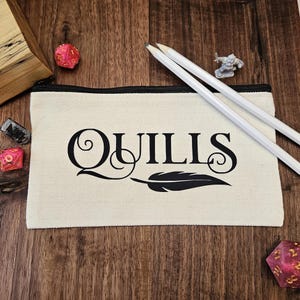 May include: A cream-colored canvas pencil case with a black zipper and the word "QUILLS" printed in black script with a feather design. Two white pencils and various gaming dice are on a wooden surface.