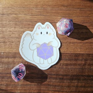May include: A white cat sticker holding a purple D20 die, with two other dice nearby. One die is a D20, and the other is a D6. The dice are pink and purple. The sticker and dice are on a wooden surface.