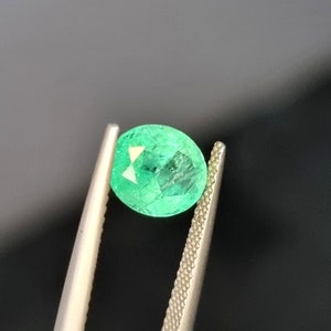 May include: A faceted, oval-shaped emerald gemstone held by tweezers. The emerald is a vibrant green colour with visible internal inclusions and a polished surface. The tweezers are silver-coloured and have textured grips.