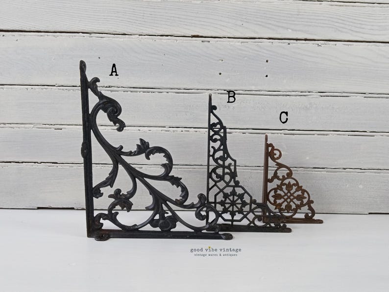 May include: Three decorative cast iron shelf brackets in varying styles. The brackets are in black and brown tones. The brackets are labeled A, B, and C. The brackets are in a vintage style.