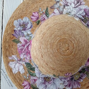 May include: A wide-brimmed sun hat with a natural woven texture, adorned with a floral design featuring purple, pink, and white peonies and butterflies. The hat has a rounded crown and a wide brim, perfect for sun protection.