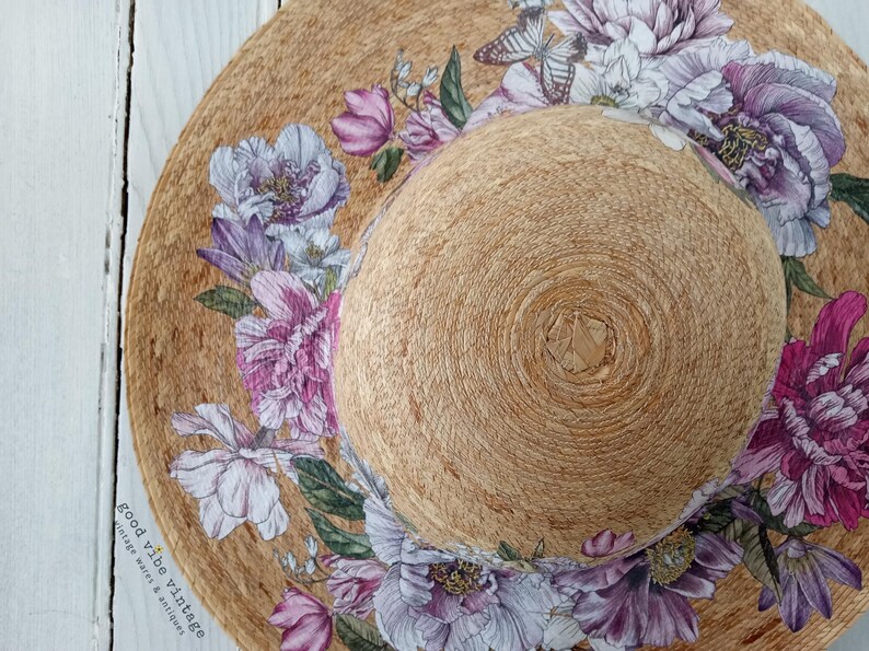 May include: A wide-brimmed sun hat with a natural woven texture, adorned with a floral design featuring purple, pink, and white peonies and butterflies. The hat has a rounded crown and a wide brim, perfect for sun protection.