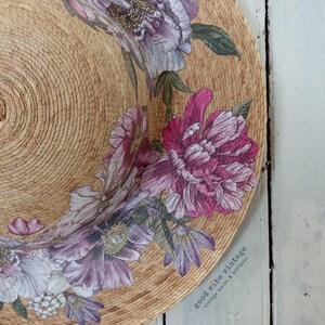 May include: A round, woven straw decorative piece with a floral design. The flowers are in shades of pink, purple, and white, with green leaves. The text "good vibe vintage" is visible on the right side.