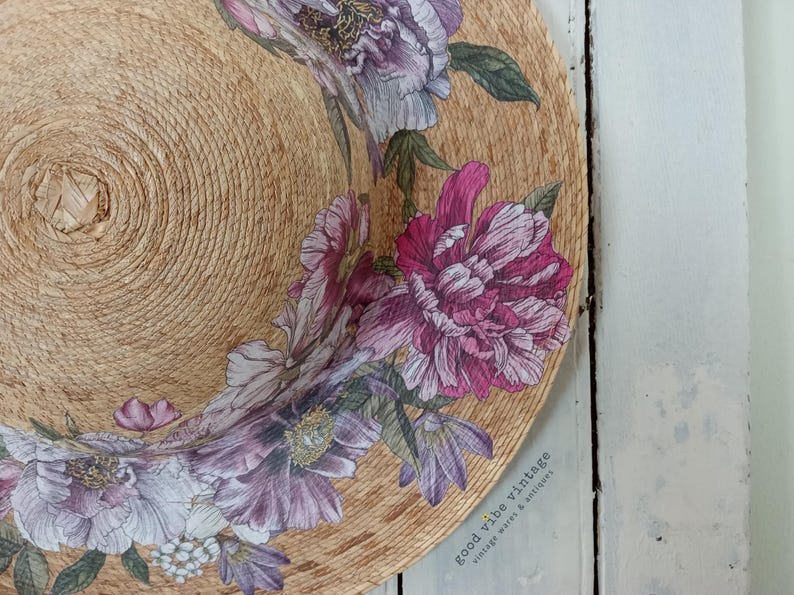 May include: A round, woven straw decorative piece with a floral design. The flowers are in shades of pink, purple, and white, with green leaves. The text "good vibe vintage" is visible on the right side.