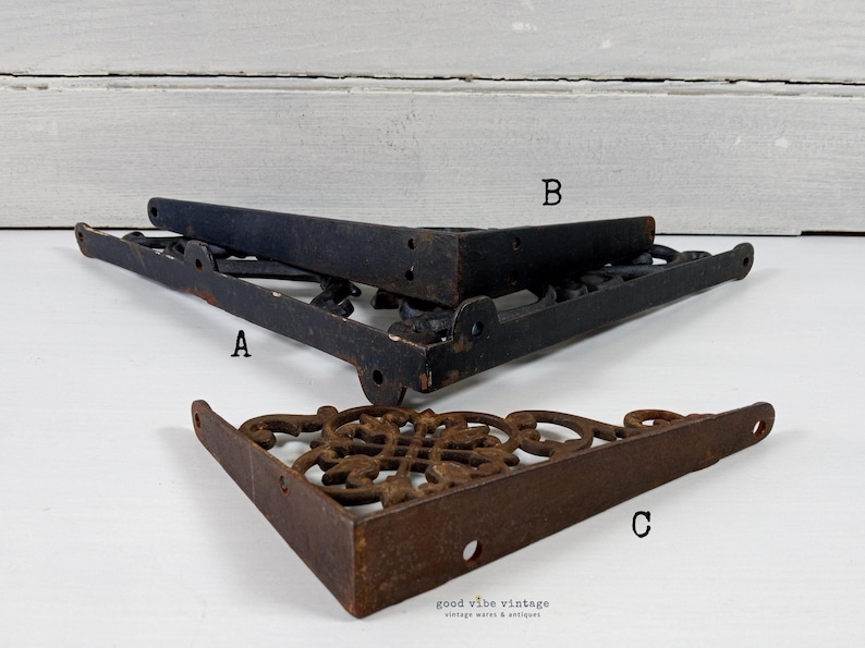 May include: Three vintage cast iron shelf brackets. Two are black, and one is a rusty brown color. The brackets are in a corner shape with decorative scrollwork. The brackets are labeled A, B, and C.