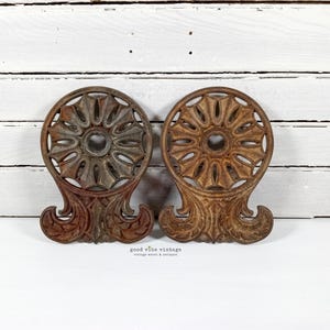 May include: Two antique cast iron decorative pieces. Each has a circular design with a central hole and ornate, scroll-like base. The pieces are in shades of brown and rust, with visible wear, suggesting age and vintage appeal.