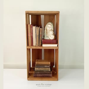 May include: A wooden crate repurposed as a bookshelf, filled with vintage books of varying sizes and colors. A statue of Mary and baby Jesus sits on the top shelf. The bottom shelf holds a stack of older books. The words "good vibe vintage" are at the bottom.