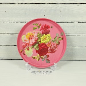 Puede incluir: Una bandeja redonda rosa con un diseño floral. La bandeja presenta un ramo de rosas en tonos rojos, amarillos y rosas. Se muestra sobre un soporte transparente contra un fondo de madera blanca. El texto "good vibe vintage" es visible.