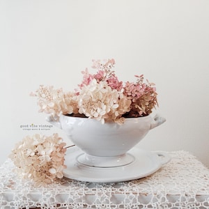 May include: A white ceramic soup tureen filled with dried pink and cream-colored flowers, resting on a matching saucer. The tureen has handles and sits on a white lace tablecloth. The words "good vibe vintage" are visible.