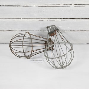 May include: Two vintage metal wire light cage shades. One shade is a rusty brown color, and the other is a silver color. The shades are on a white surface with a white wooden background.