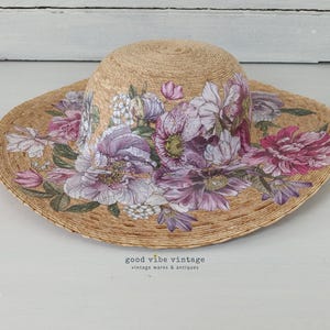 May include: A straw sun hat with a wide brim, adorned with a floral design in shades of purple, pink, and white. The hat features the text "good vibe vintage vintage wares & antiques".