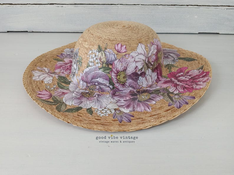 May include: A straw sun hat with a wide brim, adorned with a floral design in shades of purple, pink, and white. The hat features the text "good vibe vintage vintage wares & antiques".