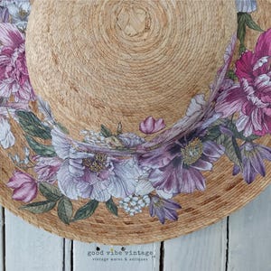 May include: A straw sun hat with a wide brim, adorned with a floral design in shades of purple, pink, and white. The hat features a natural woven texture and the text "good vibe vintage" at the bottom.