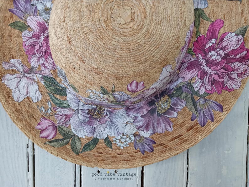 May include: A straw sun hat with a wide brim, adorned with a floral design in shades of purple, pink, and white. The hat features a natural woven texture and the text "good vibe vintage" at the bottom.